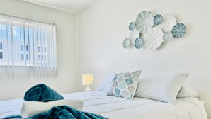 Una cama con sábanas ordenadas, una colcha blanca, un edredón color turquesa y un cojín estampado. Se observa una lámpara fijada a la pared y una ventana con persianas.