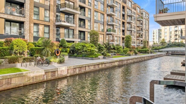 A modern apartment building with balconies, a canal, and well-maintained greenery. - Canal Side Luxury 1 Bed Chelsea (Greater London)