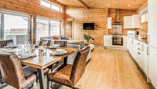 A wooden cabin interior with a dining area, sofa, kitchen, and large windows.