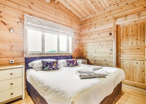 Quarto de cabana de madeira com uma cama, duas mesinhas de cabeceira, uma janela com cortinas e uma porta de madeira.