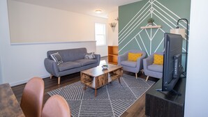 A living room with a grey sofa, two armchairs, a coffee table, a TV, and a geometric wall design.