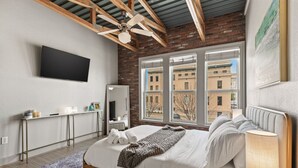 A modern bedroom with a large bed, a flat-screen TV, a window with a view of buildings, and a ceiling fan. - Intimate 2BD with Unique Style in the heart of FW (Fort Worth)