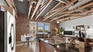 A modern kitchen with exposed beams, a brick wall, and large windows. - Intimate 2BD with Unique Style in the heart of FW (Fort Worth)