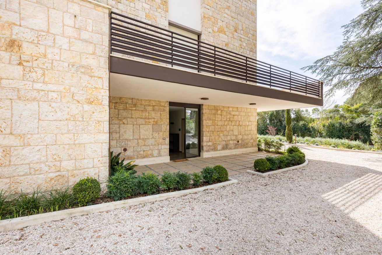 Un edificio moderno con facciata in pietra, un balcone con ringhiera in metallo e una strada di accesso sterrata.