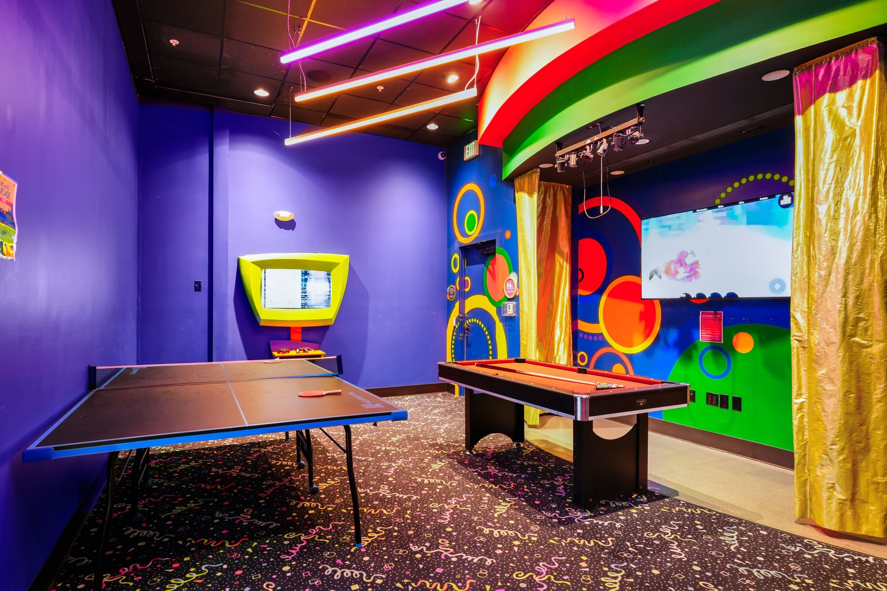A room with a ping pong table, a foosball table, and a large screen displaying a game.