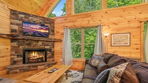 A cozy cabin interior with a stone fireplace, a wooden sofa, and a large window with curtains. - Centrally Located!~Newly Built Rustic Modern Cabin!~Guest Favorite! (Sevierville)