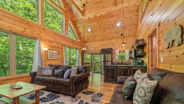 A spacious wooden cabin with a living area featuring a brown leather sofa, a wooden coffee table, and a rug. Large windows provide ample natural light and views of the surrounding forest. - Centrally Located!~Newly Built Rustic Modern Cabin!~Guest Favorite! (Sevierville)