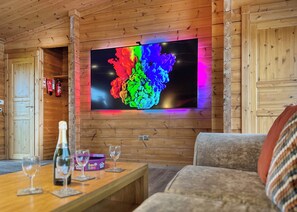 Interior de madera con un televisor de pantalla plana en la pared, una mesa de madera con vasos y una botella, y un sofá en primer plano.