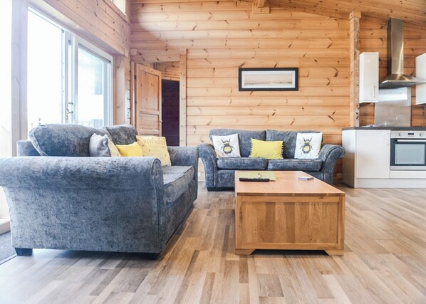 A log cabin interior with two grey sofas, a wooden coffee table, and a kitchen area.