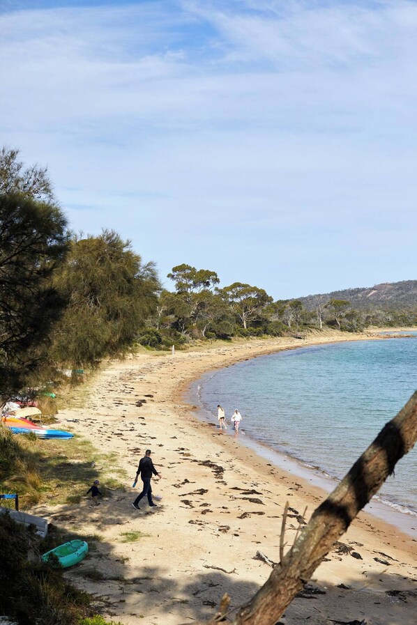 A sandy beach with a few people walking, a clear sky, and trees in the background.