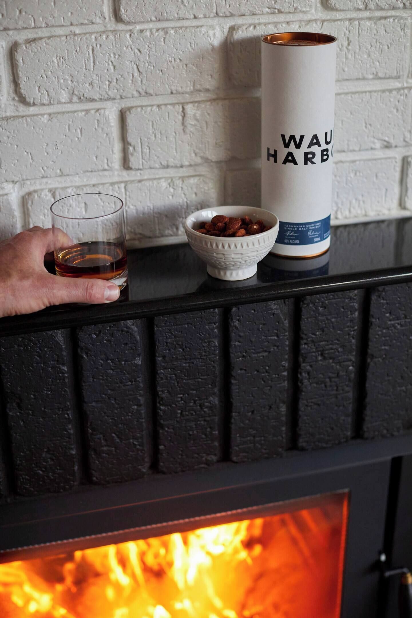 En hand som håller i ett glas whisky, en skål med nötter och ett ljus med etiketten ”Wau Harbo” placerat på en hylla ovanför en öppen spis.