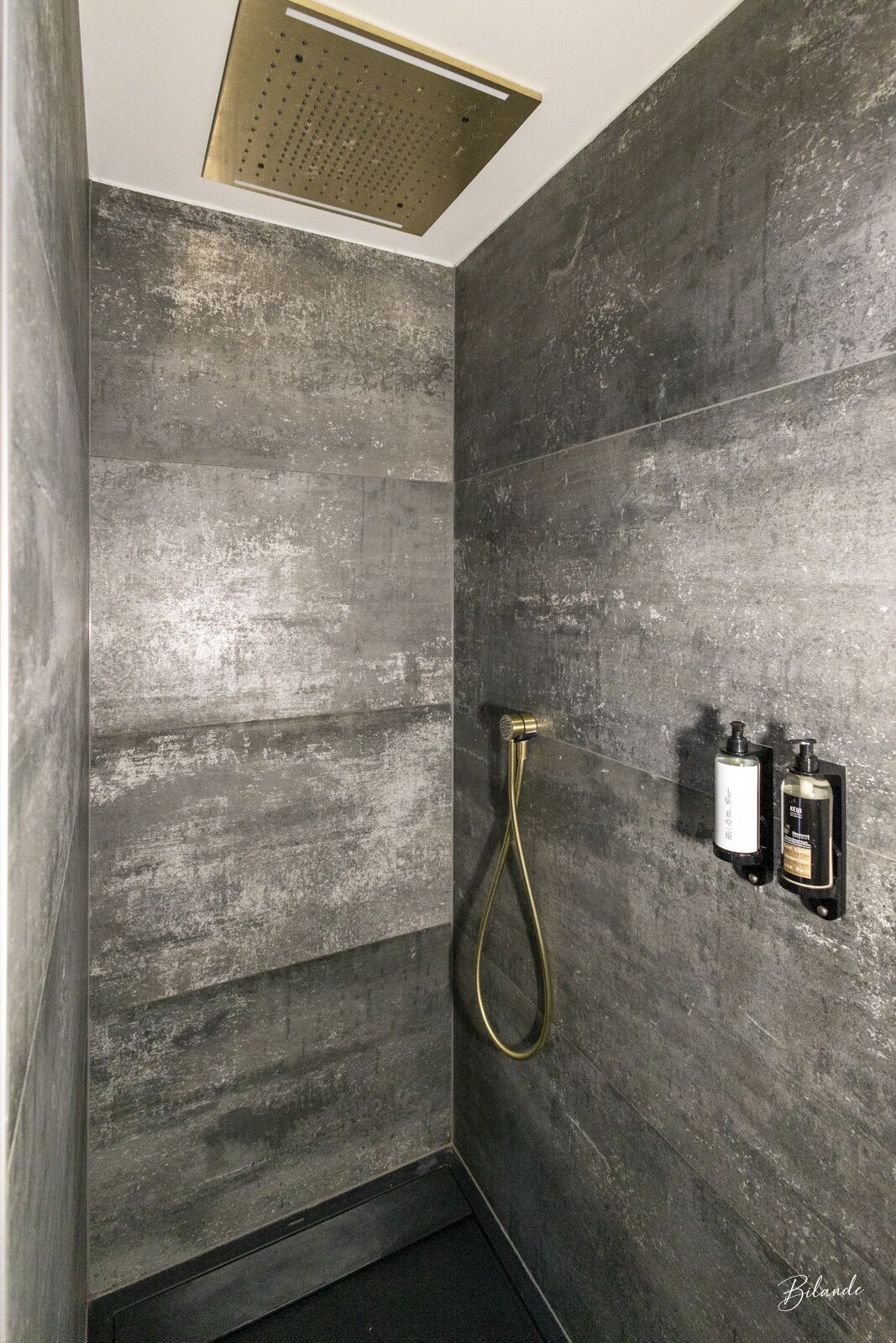 A modern bathroom with dark stone walls, a shower with a gold showerhead, and two wall-mounted soap dispensers.