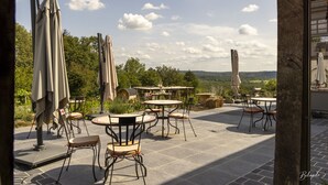 An outdoor patio with round tables and chairs, surrounded by umbrellas, set against a backdrop of trees and a distant view. - La chocolaterie de Roly (Philippeville)