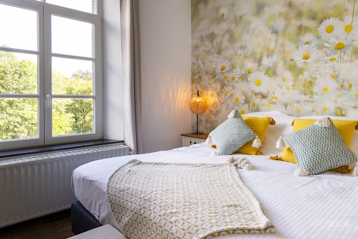 A bedroom with a large window, a bed with white bedding, and a wall decorated with a daisy pattern.