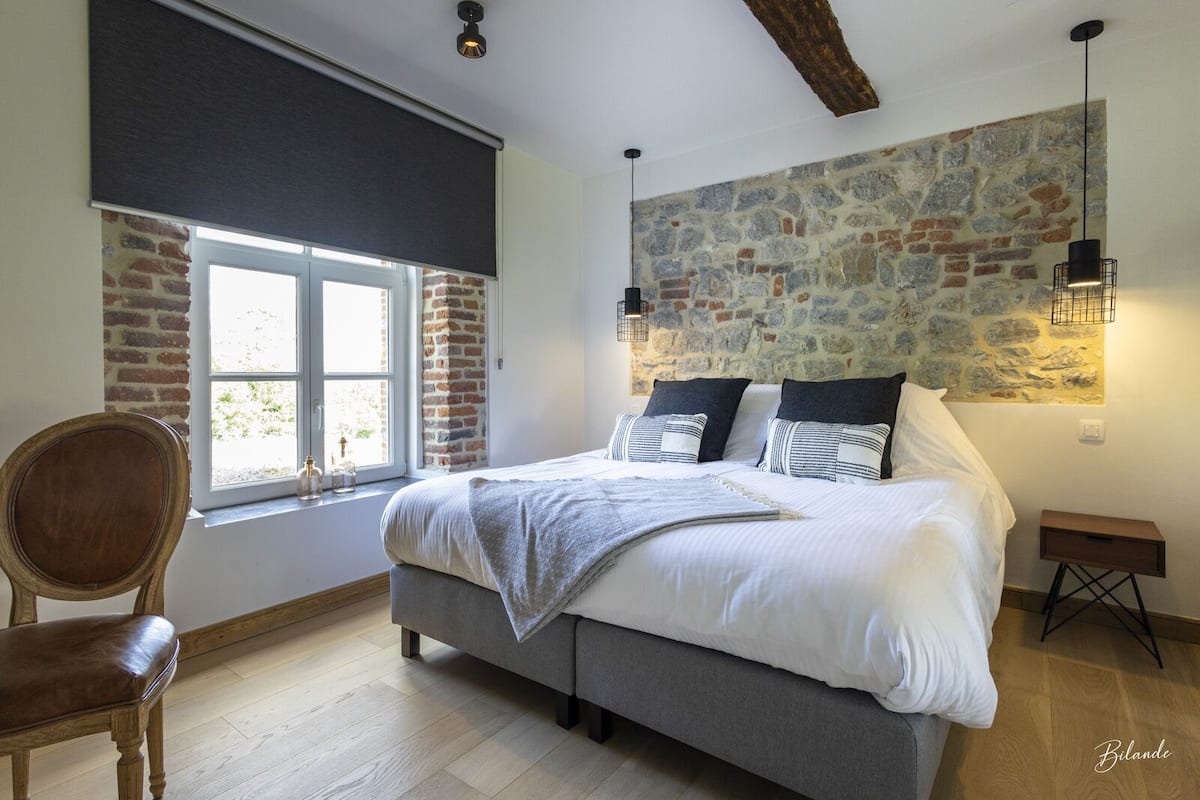 A bedroom with a large bed, a chair, a window with a view, and a stone wall.