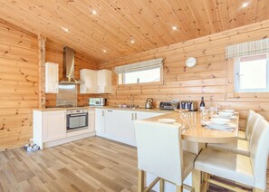 A wooden cabin interior with a kitchen and dining area, featuring wooden walls, a stove, cabinets, and a dining table set for four.