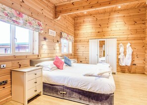 A wooden cabin bedroom with a bed, bedside tables, a dresser, a window with a floral curtain, and a wardrobe.
