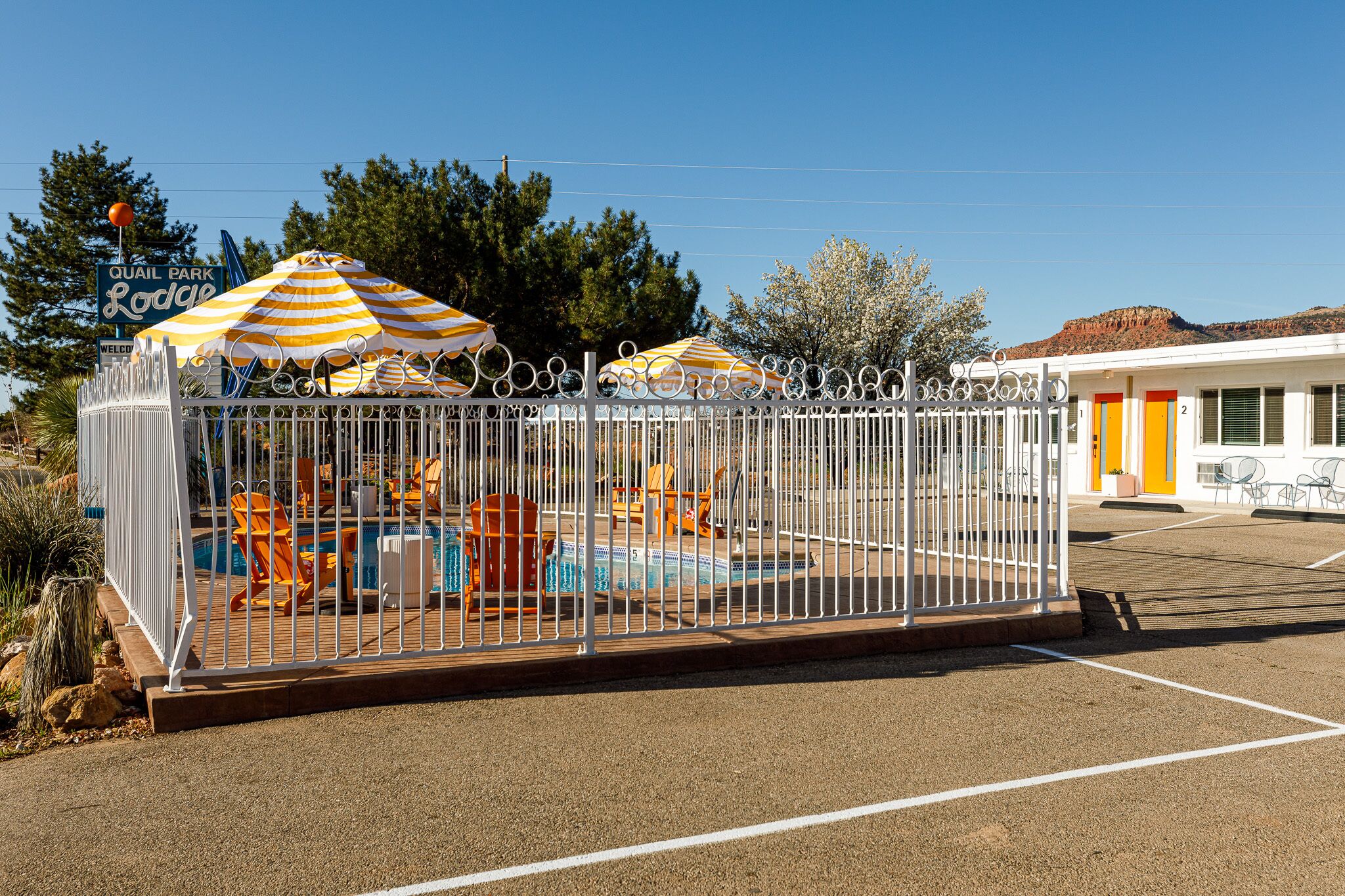 Zona de piscina con valla, sombrillas a rayas, sillas de descanso y un edificio con puerta amarilla.