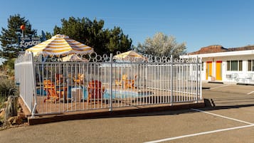 Zona de piscina con valla, sombrillas a rayas, sillas de descanso y un edificio con puerta amarilla.