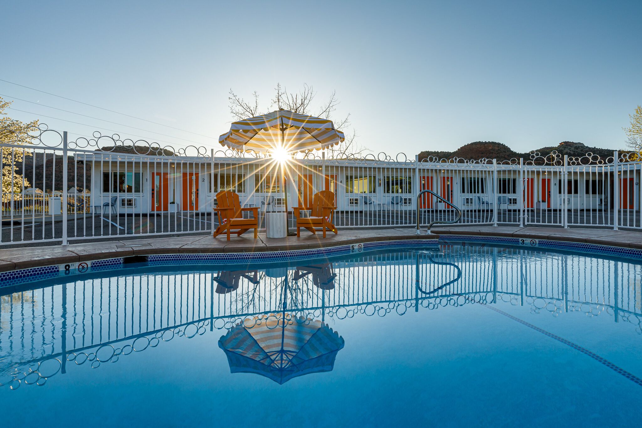 A swimming pool with a sunlit umbrella, a fenced area with orange chairs, and a view of a mountainous landscape.