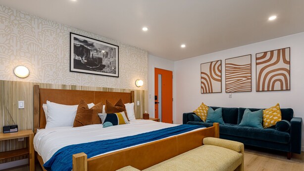 Una habitación de hotel moderna con una cama grande, un sofá y un banco. Hay luces fijadas a la pared y un cuadro enmarcado sobre la cama.