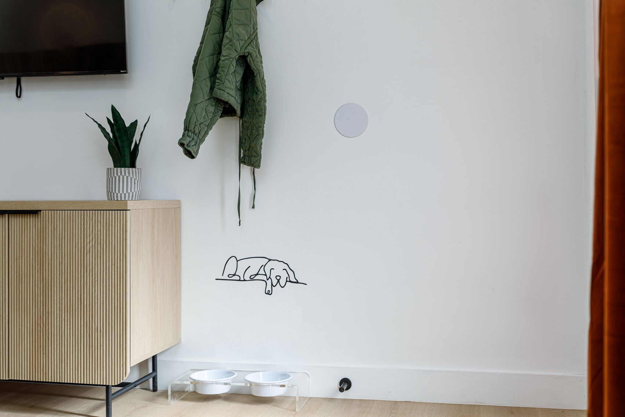 A room with a wooden cabinet, a green jacket hanging, a potted plant, a wall decal of a sleeping dog, and two pet bowls on the floor.
