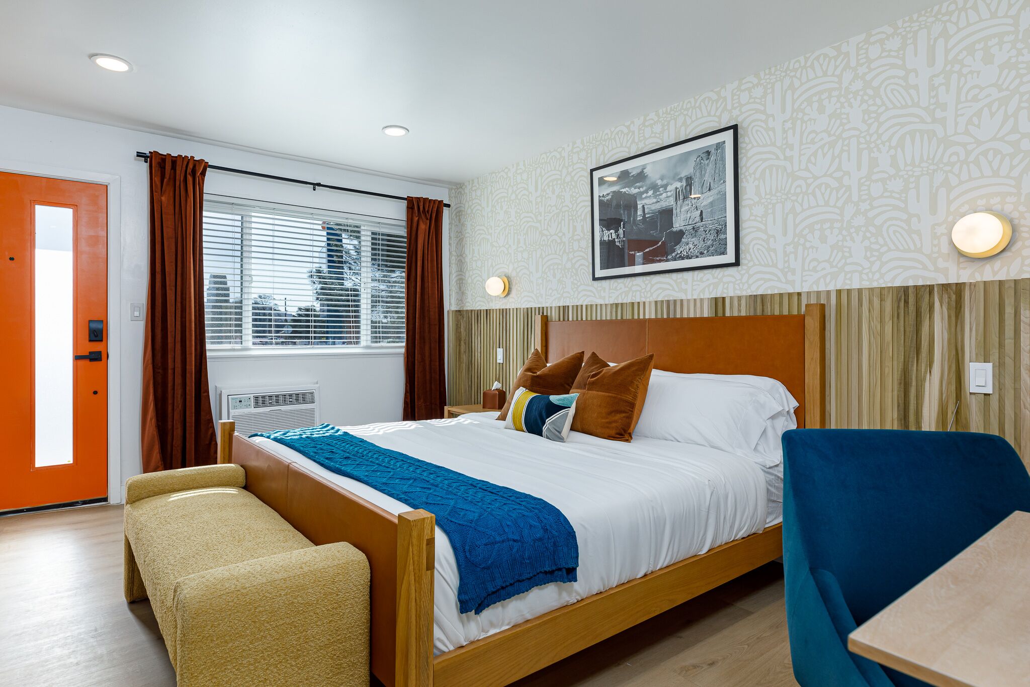 A hotel room with a bed, a bench, a table, a window with curtains, a wall-mounted picture, and a patterned wallpaper.