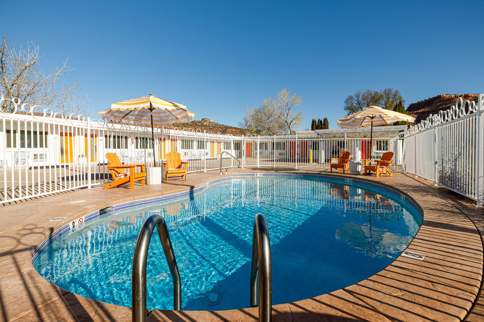 A swimming pool with a wooden deck, surrounded by a white fence and orange chairs.