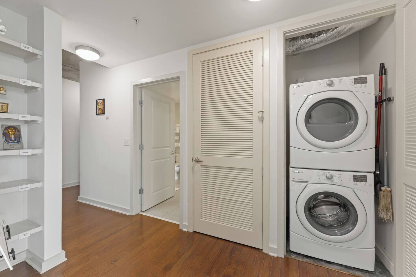 Un lavadero con dos lavadoras y secadoras apiladas, un escoba y una puerta que da a otra habitación.