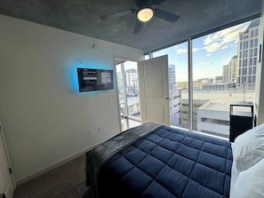 A hotel room with a bed, a ceiling fan, a mounted TV, and a view of the cityscape through large windows.