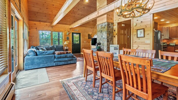 A spacious log cabin interior with a dining area, a stone fireplace, and a large window overlooking the forest. - Whippoorwill - Wooded location, hot tub, close to downtown Oquossoc (Rangeley)
