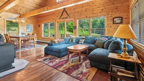 A cozy cabin interior with a wooden ceiling, a blue sofa, a dining table, and a large window. - Whippoorwill - Wooded location, hot tub, close to downtown Oquossoc (Rangeley)