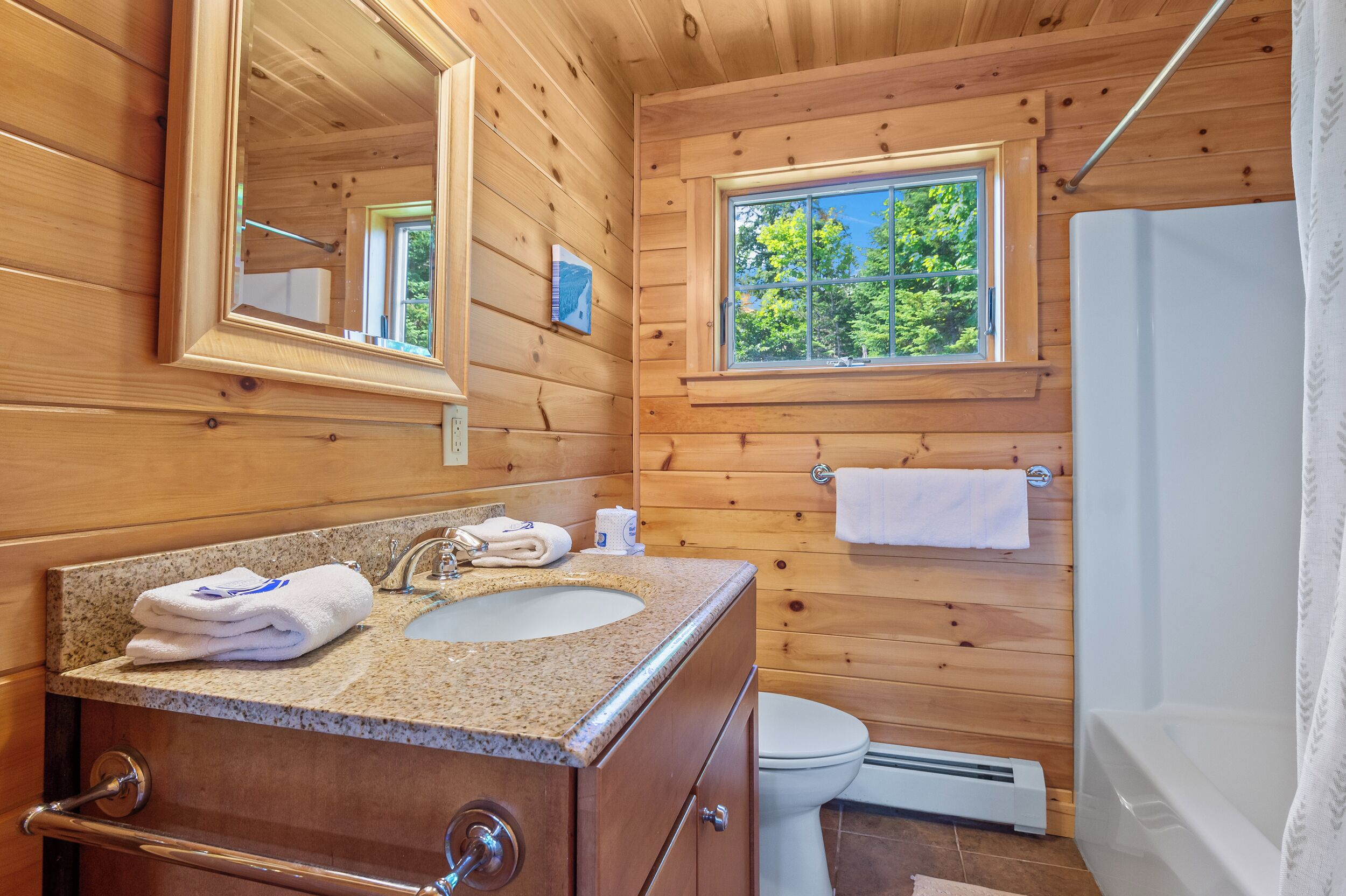 Ein Badezimmer einer Holzhütte mit einem Granittisch, einem Spiegel, einem Fenster, einer Toilette und einer Badewanne.