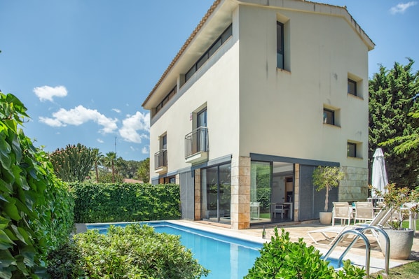 Un edificio de dos pisos con piscina, mobiliario exterior y vista a la vegetación circundante.