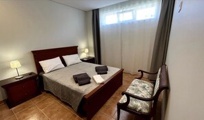 A neatly made bed with pillows and folded towels, a bedside lamp, a wooden nightstand, and a chair with a patterned cushion.