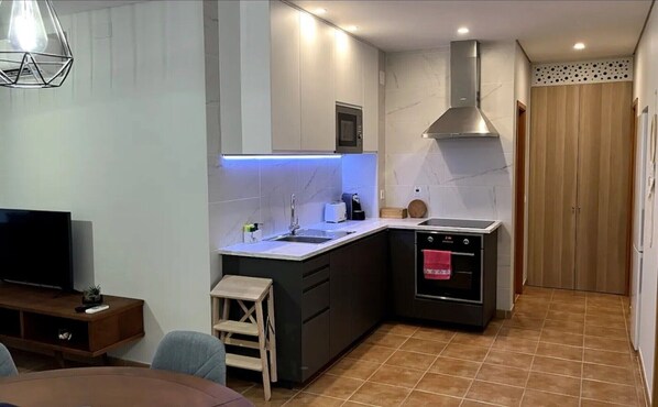 A compact kitchen with a microwave, stove, and sink, a small wooden stool, and a TV in the corner.