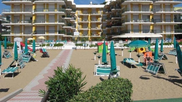 Hotel com vários andares, varandas, área de praia com espreguiçadeiras e guarda-sóis, e um céu azul límpido.