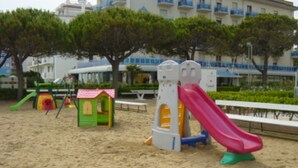 Um parquinho de areia com um escorrega, uma casinha de brincar verde e bancos.