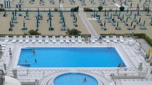Uma piscina grande e retangular com uma piscina circular menor no centro, rodeada por espreguiçadeiras e guarda-sóis.