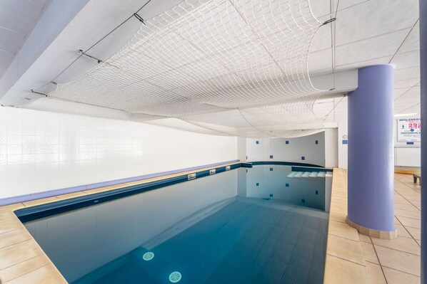 Une piscine intérieure avec un sol carrelé, un plafond en treillis et un bord de piscine bleu.