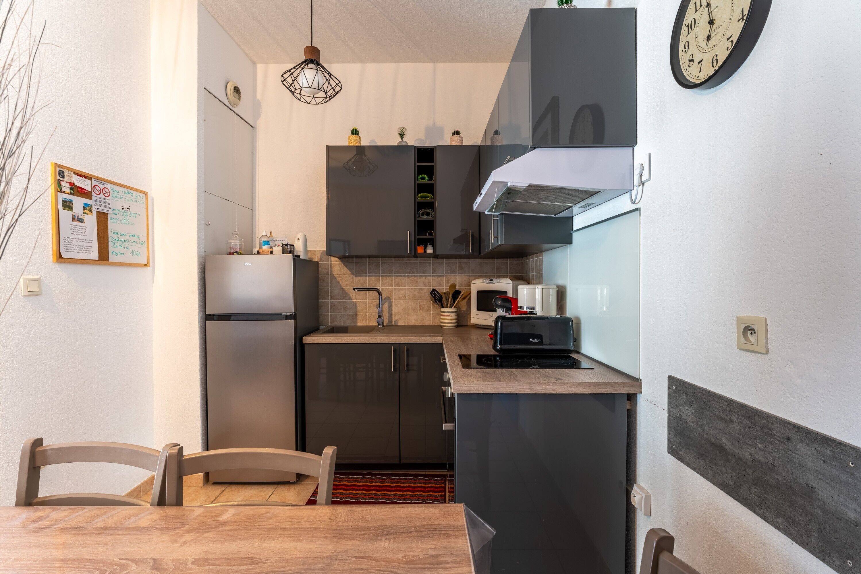 Moderni keittiö, jossa on sisäänrakennettu uuni, mikroaaltouuni ja kahvinkeitin, sekä puinen ruokapöytä tuoleineen ja seinälle kiinnitetty kello.