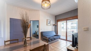 A dining area with a wooden table and chairs, a vase of dried branches, a sofa, a television, and a sliding glass door leading to a balcony. - Apartment 'Appartement Des Cedres' with Shared Pool, Wi-Fi and Air Conditioning (Uzès)