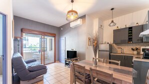 A modern living room with a sofa, dining table, chairs, a TV, and a kitchen area with cabinets and a sink. - Apartment 'Appartement Des Cedres' with Shared Pool, Wi-Fi and Air Conditioning (Uzès)