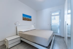 Une chambre à coucher comprenant un lit, des tables de chevet, une lampe fixée au mur, une fenêtre avec des stores et un tableau.