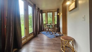 A room with large windows, a dining table with chairs, a small stool, and a wall-mounted light fixture. - Peaceful AuSable River Retreat-Frederic, MI Sleeps 6 (Frederic)
