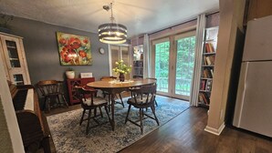 A dining area with a round table, chairs, a chandelier, a painting, and a view through a glass door. - Peaceful AuSable River Retreat-Frederic, MI Sleeps 6 (Frederic)