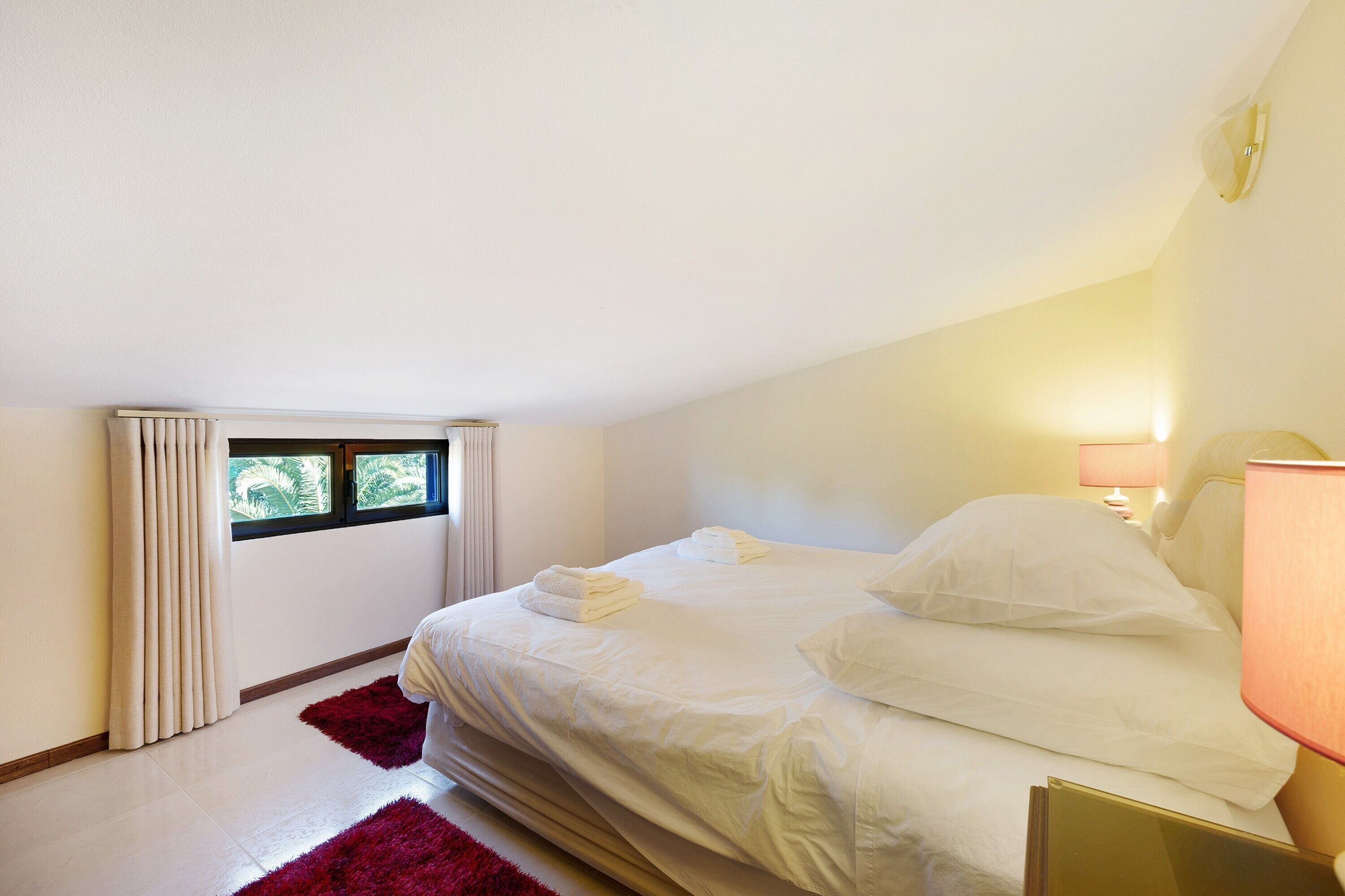 Een slaapkamer met een bed, een raam, een lamp en een rood tapijt.