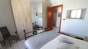A bedroom with a bed, a chair, a wooden wardrobe, a mirror, and a door leading to another room. - Surya's House Otranto (Otranto)