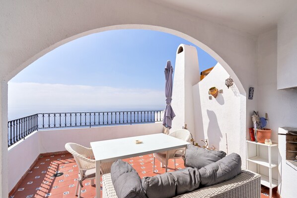 A balcony with a white table, wicker chairs, grey cushions, a white shelf, and a view of the sea.