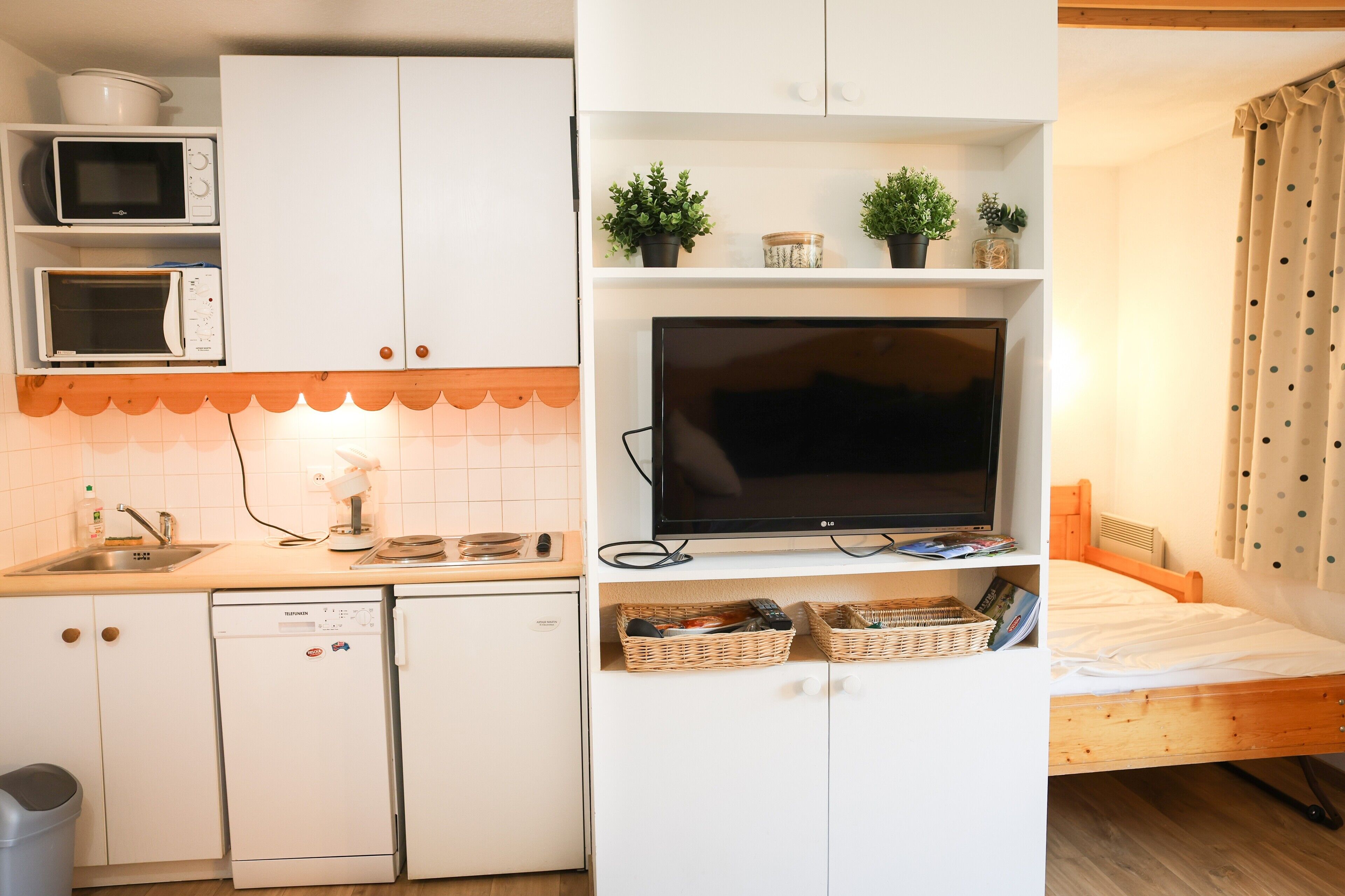 A compact kitchen with a microwave, toaster, and dishwasher, a flat-screen TV, and a bed with a wooden headboard.